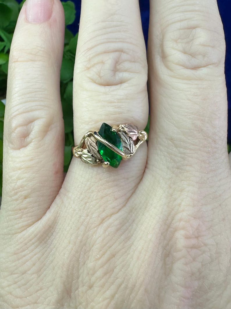 May include: A gold ring featuring a green gemstone and leaf accents. The ring is worn on a finger.