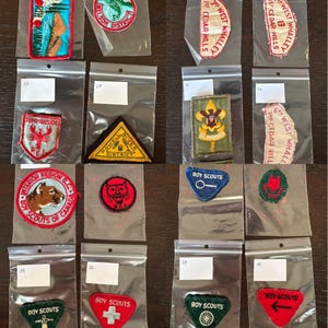 May include: A collection of 10 Boy Scout badges in plastic bags. The badges are in various colours and designs, including a red badge with a white cross, a blue badge with a key, and a green badge with a ship's wheel. The badges are all in good condition.
