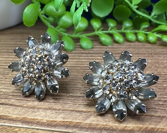 Op de afbeelding: Een paar zilverkleurige oorbellen met een bloemmotief. Elke oorbel heeft in het midden een cluster van heldere strass steentjes, omgeven door zilveren bloemblaadjes.