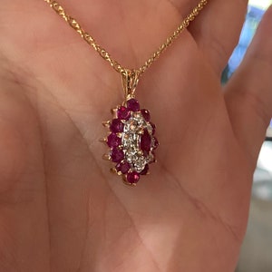 Op de afbeelding: Een gouden ketting met een ovaalvormige hanger. De hanger is bezet met een centrale marquise-geslepen rode edelsteen, omringd door een cluster van kleinere ronde rode edelstenen en kleine heldere steentjes in een bloemachtig patroon.