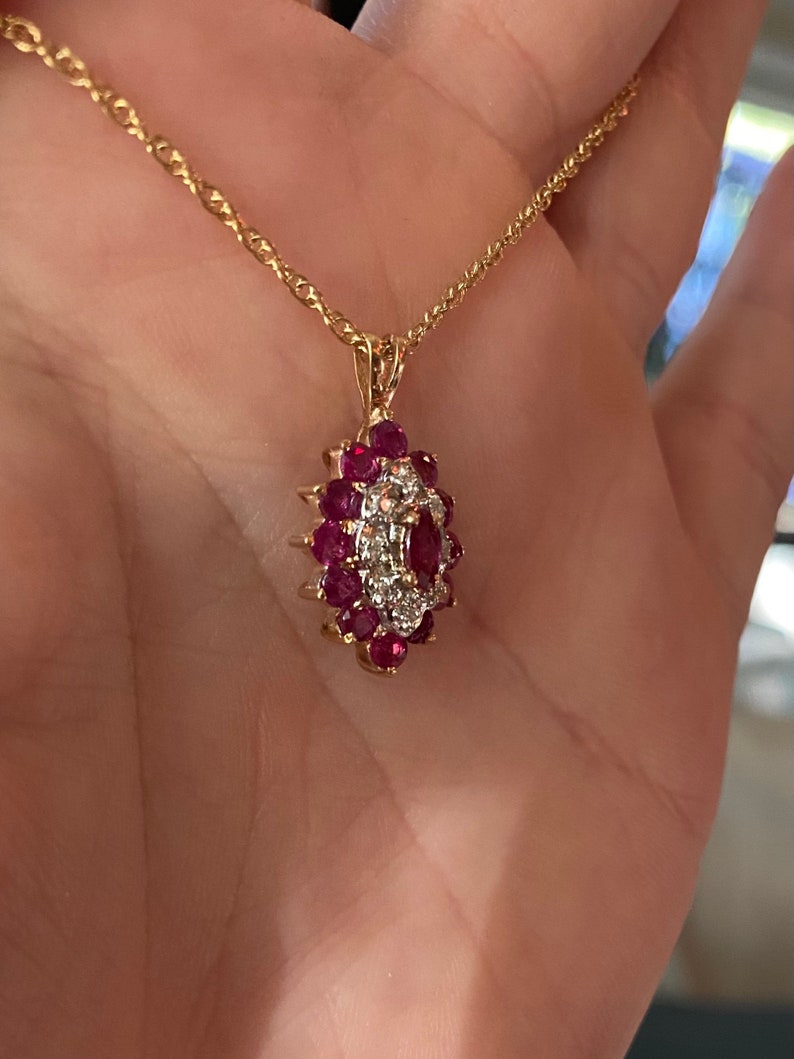 Op de afbeelding: Een gouden ketting met een ovaalvormige hanger. De hanger is bezet met een centrale marquise-geslepen rode edelsteen, omringd door een cluster van kleinere ronde rode edelstenen en kleine heldere steentjes in een bloemachtig patroon.