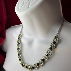 May include: A silver-toned necklace with a floral design. The necklace features small, dark blue and iridescent gemstones set within the flower-shaped links. The necklace is displayed on a white mannequin.