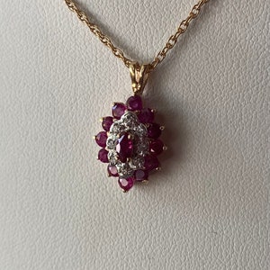 Op de afbeelding: Een gouden ketting met een hanger voorzien van een centrale robijn in marquise-slijpvorm, omringd door een halo van kleine witte diamanten, die weer is omcirkeld door een cluster van ronde robijnen in een bloemvormig ontwerp op goud.