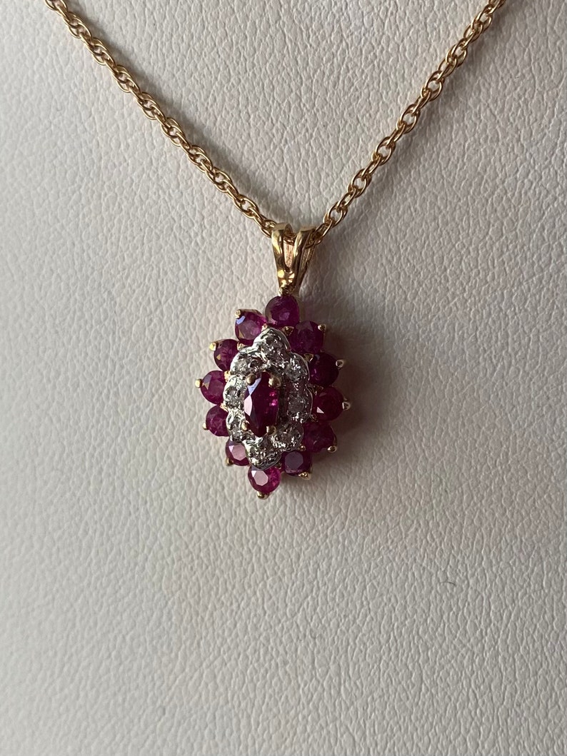 Op de afbeelding: Een gouden ketting met een hanger voorzien van een centrale robijn in marquise-slijpvorm, omringd door een halo van kleine witte diamanten, die weer is omcirkeld door een cluster van ronde robijnen in een bloemvormig ontwerp op goud.