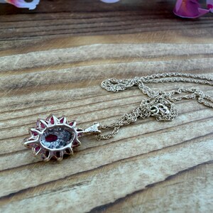Op de afbeelding: Een zilverkleurige ketting met een ovaalvormige hanger met een dieprode steen in het midden, omringd door een halo van kleine, heldere, fonkelende steentjes. De hanger zit vast aan een fijne, getextureerde zilverkleurige ketting met een sluiting.