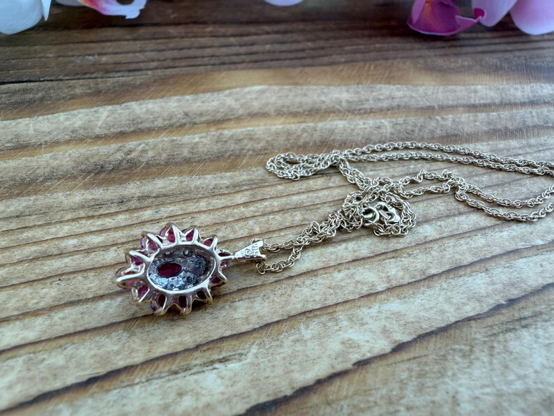 Op de afbeelding: Een zilverkleurige ketting met een ovaalvormige hanger met een dieprode steen in het midden, omringd door een halo van kleine, heldere, fonkelende steentjes. De hanger zit vast aan een fijne, getextureerde zilverkleurige ketting met een sluiting.