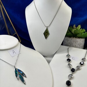 May include: Three silver necklaces with different pendants. The first necklace has a green and blue abalone shell pendant. The second necklace has a diamond-shaped pendant with a green and blue abalone shell. The third necklace has a silver chain with black gemstone pendants.