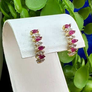 May include: A pair of gold hoop earrings featuring a line of marquise-shaped red and white gemstones. The earrings are shown on a white jewellery stand.