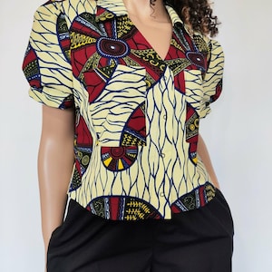May include: A short-sleeved shirt with a collar and an African-inspired print. The shirt has a V-neck and a colourful pattern of red, yellow, blue, and black geometric shapes on a cream background. It is worn with black trousers.