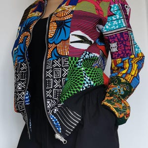 Op de afbeelding: Een bomberjack met lange mouwen voorzien van een levendig patchworkontwerp met diverse kleurrijke, op Afrika geïnspireerde prints, waaronder geometrische patronen, bloemmotieven en een vogelafbeelding, afgewerkt met een zilverkleurige rits.