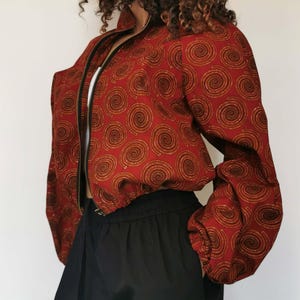 Könnte beinhalten: Eine bauchfreie Bomberjacke in Dunkelrot mit einem sich wiederholenden Muster aus goldbraunen Spiralen. Die Jacke hat einen schwarzen Frontreißverschluss, einen Stehkragen und lange Ärmel mit gerafften elastischen Bündchen, kombiniert mit einer schwarzen Hose.