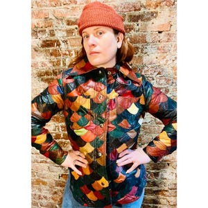 May include: A long-sleeve button-up jacket featuring a multi-colored scalloped pattern in shades of red, orange, yellow, green, and black. The jacket has a pointed collar, two chest pockets, and metallic buttons. A solid orange knit beanie is worn.