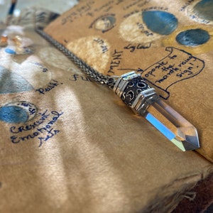 Op de afbeelding: Een zilveren ketting met een kristallen hanger en een gedetailleerde zilveren bedel. De ketting ligt op een stuk papier met handgeschreven notities over de maan en astrologie.