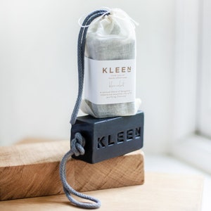 Può includere: Una saponetta nera con la scritta "KLEEN" in rilievo. Il sapone è avvolto in un sacchetto di mussola bianca con un cordoncino di chiusura grigio. Il sacchetto ha un'etichetta che recita "KLEEN Sapone artigianale 100% naturale Blue Velvet Un mix sensuale di bergamotto e oli essenziali di legno di cedro con carbone purificante."