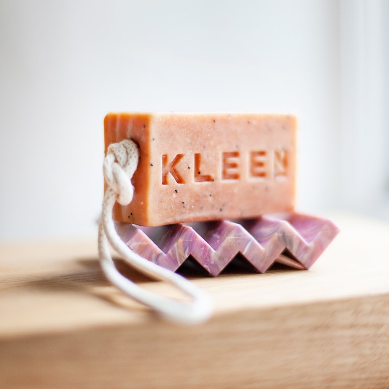 May include: A pink bar of soap with the word "KLEEN" embossed on it. The soap is on a purple and white soap dish with a white rope attached to it.