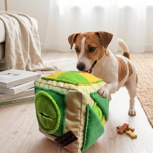 May include: A soft, cube-shaped dog puzzle toy featuring a light tan base with bright green and yellow felt accents. The toy includes a circular green pocket with a slit opening and triangular green flaps, designed for hiding small treats.