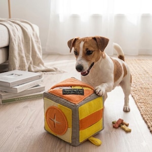 May include: A soft, fabric cube toy for pets featuring yellow and orange panels, a circular orange applique with a cross-shaped cutout, and a black label with the text "Togee Dog Toys homemade" on the top edge, with a small dog nearby.