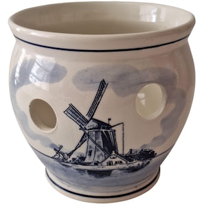 May include: A white ceramic onion keeper with a blue windmill design. The round container has two large holes and a blue rim. The windmill scene is painted in blue, with a water scene at the bottom.