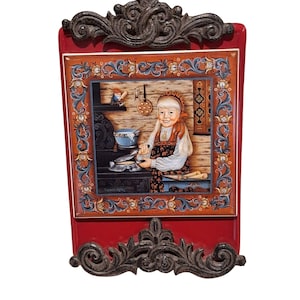May include: A rectangular, red decorative plaque with a detailed painting of a young girl cooking in a kitchen. The painting is framed with ornate floral designs and a dark, decorative border. The plaque has a dark, decorative top and bottom.