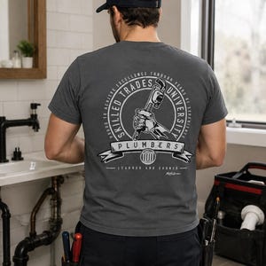 May include: A dark gray t-shirt featuring a circular graphic on the back with a hand holding a pipe wrench. The text reads Skilled Trades University, Plumbers, and Learned and Earned. The shirt is paired with black work pants and a navy blue baseball cap.