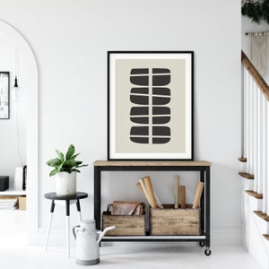 May include: Black and white abstract art print in a black frame, hanging on a white wall above a wooden shelf with black metal legs.