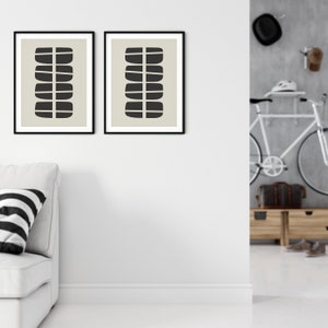 May include: Two black framed prints featuring abstract geometric shapes in black against a beige background. The prints are hung on a white wall above a white sofa with a black and white striped cushion.
