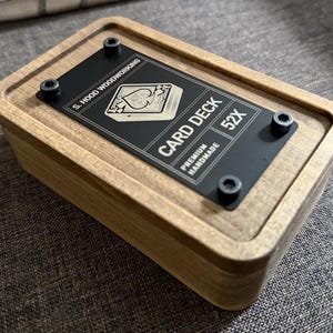 May include: A wooden card deck box with a black metal plate on top. The plate is secured with four screws and features the text "CARD DECK 52X" and "PREMIUM HANDMADE". The box is made of light-colored wood.