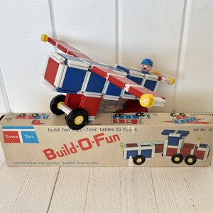 Puede incluir: Un juego de juguetes Build-O-Fun vintage con un avión rojo, blanco y azul y una caja con el texto "Build-O-Fun". El avión de juguete tiene una pequeña figura de piloto y ruedas negras. La caja también muestra un tren.