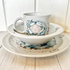 Op de afbeelding: Een witte keramische kinderset met een blauw en wit bloemenmotief. De set bevat een kom, een bord en een mok met een cartoon gezicht.