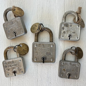 Puede incluir: Cinco candados Master vintage con llaves. Los candados son plateados, cuadrados y tienen asas arqueadas. Cada candado tiene una llave y una pequeña etiqueta redonda de latón. Están sobre una superficie de madera blanca.