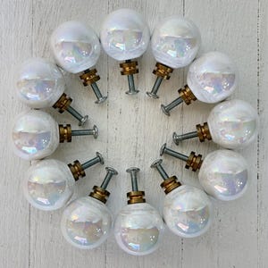Peut inclure: Une collection de douze boutons de placard en verre blanc irisé disposés en cercle. Chaque bouton a une base en laiton et une vis argentée. Les boutons sont posés sur un fond en bois blanc.