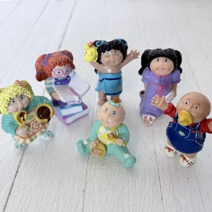 Può includere: Una collezione di sei piccole statuette in plastica Cabbage Patch Kids. Le figure includono un bambino biondo con un cucchiaio, un bambino dai capelli rossi che legge un libro, un bambino con un asciugamano blu, un bambino con un vestito viola, un neonato con bavaglino e un neonato calvo con salopette.