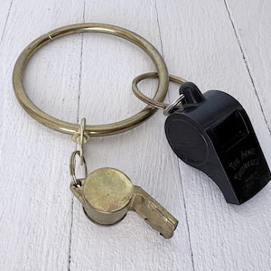 Puede incluir: Una colección de silbatos y llaveros. La imagen presenta un gran llavero de latón con un pequeño silbato de latón y un silbato de plástico negro adjunto. El silbato negro tiene el texto "The Acme Thunderer" impreso.