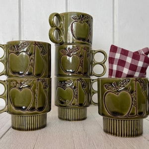 Könnte beinhalten: Ein Set aus sechs olivgrünen Keramikbechern mit einzigartigen Doppel-Schlaufen-Griffen. Jeder Becher zeigt geprägte Obstmuster von Äpfeln und Birnen. Die Becher sind gestapelt und auf einer weißen Oberfläche angeordnet, mit einer rot-weiß karierten Serviette.