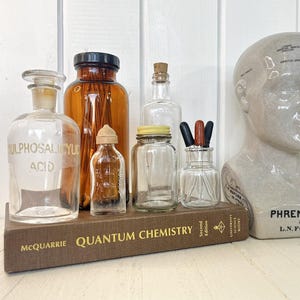 May include: A selection of antique glass bottles and jars, in amber and clear glass, with different stoppers and lids. A ceramic bust marked "Phrenology" is also visible, all displayed on a brown book titled "Quantum Chemistry".