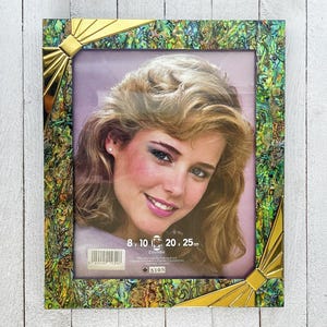 Può includere: Una cornice decorativa con un ritratto di donna. La cornice ha un design in abalone oro e verde con accenti dorati. Le dimensioni della cornice sono 20,3 x 51,4 cm.