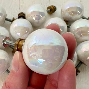 Peut inclure: Ensemble de boutons de meubles en céramique blanche irisée avec des fixations en laiton. Les boutons sphériques présentent une finition nacrée, reflétant un arc-en-ciel de couleurs. Chaque bouton comprend une tige filetée et une collerette en laiton.
