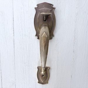 Peut inclure: Une poignée de porte verticale en laiton dotée d'une prise texturée avec un accent central en forme de losange. La poignée est montée sur deux plaques décoratives avec un motif quadrillé en haut et en bas, dans un ton métallique mat.