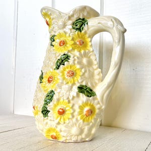 Puede incluir: Una jarra de cerámica blanca con un diseño floral. La jarra está decorada con margaritas amarillas y naranjas y hojas verdes.