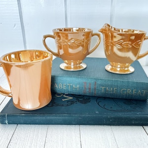 Puede incluir: Tres tazas de té y una crema de vidrio naranja iridiscente se colocan encima de dos libros azules vintage. Las tazas tienen asas y un diseño decorativo de hojas. La crema tiene un pico.