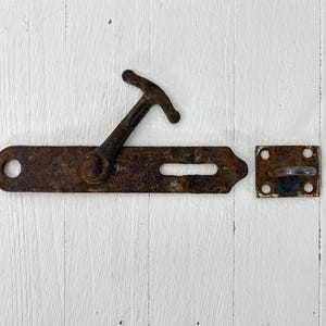 May include: A rusty metal latch and catch set against a white wooden background. The latch has a handle and a long, rectangular plate with a slot. The catch is a small square plate with a curved handle.
