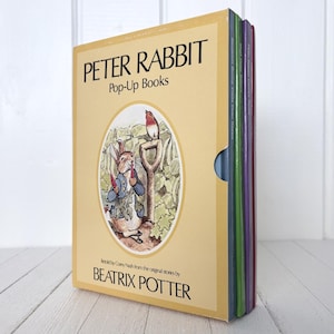 Op de afbeelding: Een boxset met Peter Rabbit pop-upboeken van Beatrix Potter. De gele cassette bevat een illustratie van Peter Rabbit en een roodborstje in een tuin, met de titel en auteur in zwarte letters. De set bevat verschillende kleurrijke boeken.