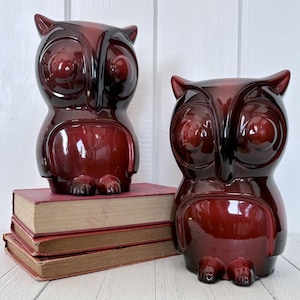 May include: Two burgundy owl ornaments with large, round eyes. One owl sits on a stack of vintage books with red covers. The owls have a glossy finish and are on a white surface.