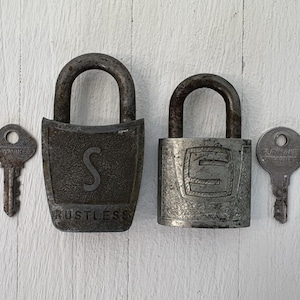 Puede incluir: Dos candados antiguos con llaves. Un candado tiene un diseño de "S" y la palabra "RUSTLESS". El otro candado tiene un diseño de "E". Tanto los candados como las llaves son de metal y muestran signos de edad.