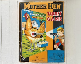 May include: Vintage "Mother Hen Target Game" box. The box features a cartoon illustration of a hen with a target on its side, a boy aiming a toy gun, and the text "Mother Hen Target Game". The box is orange, blue, and yellow.
