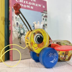 May include: A vintage wooden pull-along toy bee, yellow with blue wheels. The bee has a smiling face, a white crown, and a red propeller. A yellow string is attached. Books are in the background.