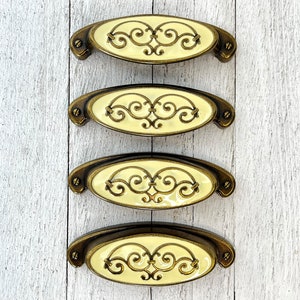 Pode incluir: Quatro botões de armário de latão antigo com um centro ovalado de cor creme com um design decorativo de rolagem.