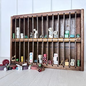 Peut inclure: Un présentoir rectangulaire en bois doté de deux rangées horizontales de compartiments verticaux. Les compartiments contiennent divers dés, petites figurines et billes. D'autres pièces de jeu, dont des jetons ronds, sont éparpillées devant.