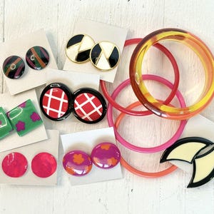 Puede incluir: Una colección de joyas vintage coloridas, que incluye pendientes y pulseras. Los pendientes presentan varios diseños, como rayas, patrones geométricos y motivos florales. Las pulseras son en tonos rosa y ámbar.