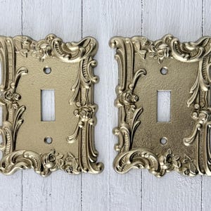 Puede incluir: Dos cubiertas de interruptor de luz doradas y ornamentadas. Cada cubierta tiene una abertura rectangular para el interruptor y está rodeada por un diseño decorativo floral y de volutas. Las cubiertas se muestran sobre un fondo de madera blanca desgastada.
